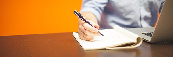 Photo of a person writing on a notebook while sitting in front of a laptop.